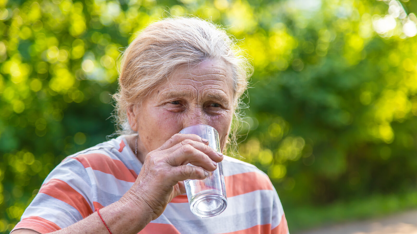 Une personne âgée qui boit un verre d’eau dans un salon aux volets mi-clos, lumière douce